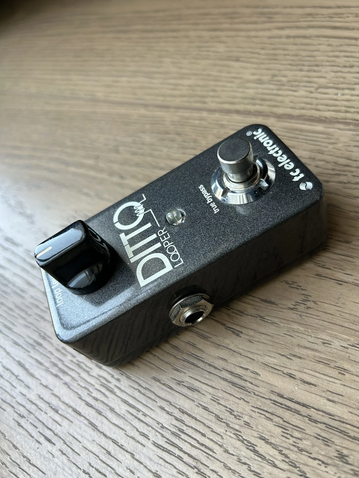 Fotografia original do pedal TC Electronic Ditto Looper sobre uma mesa de madeira, com o controle de volume, o footswitch e o conector lateral visíveis