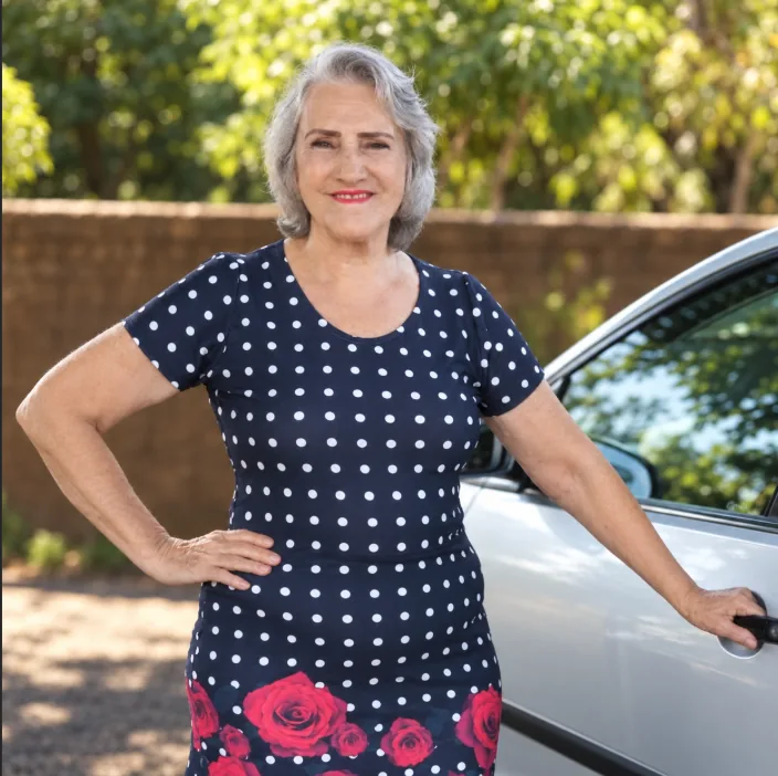Dona Zulmira, de vestido azul com bolinhas brancas e flores vermelhas, em pé ao lado de um carro.