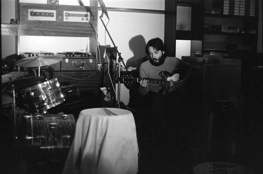 Paul McCartney sentado com uma guitarra, ao lado de uma bateria e de equipamento de gravação