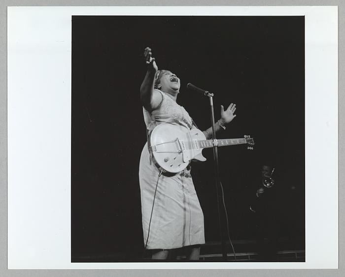 Sister Rosetta Tharpe canta diante de um microfone, com os braços erguidos e uma guitarra a tiracolo, em fotografia em preto e branco