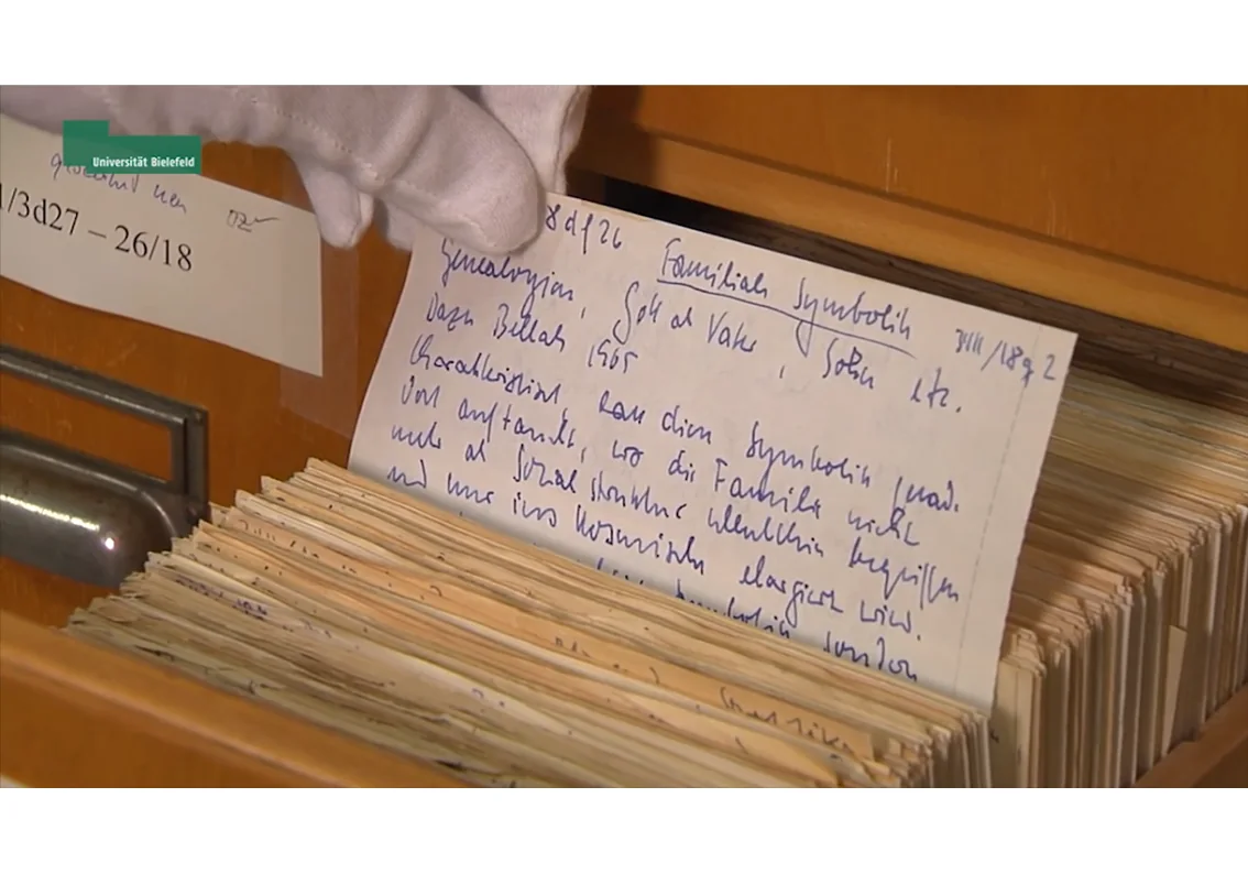 Mão enluvada segura uma ficha manuscrita diante de uma gaveta do Zettelkasten de Niklas Luhmann.